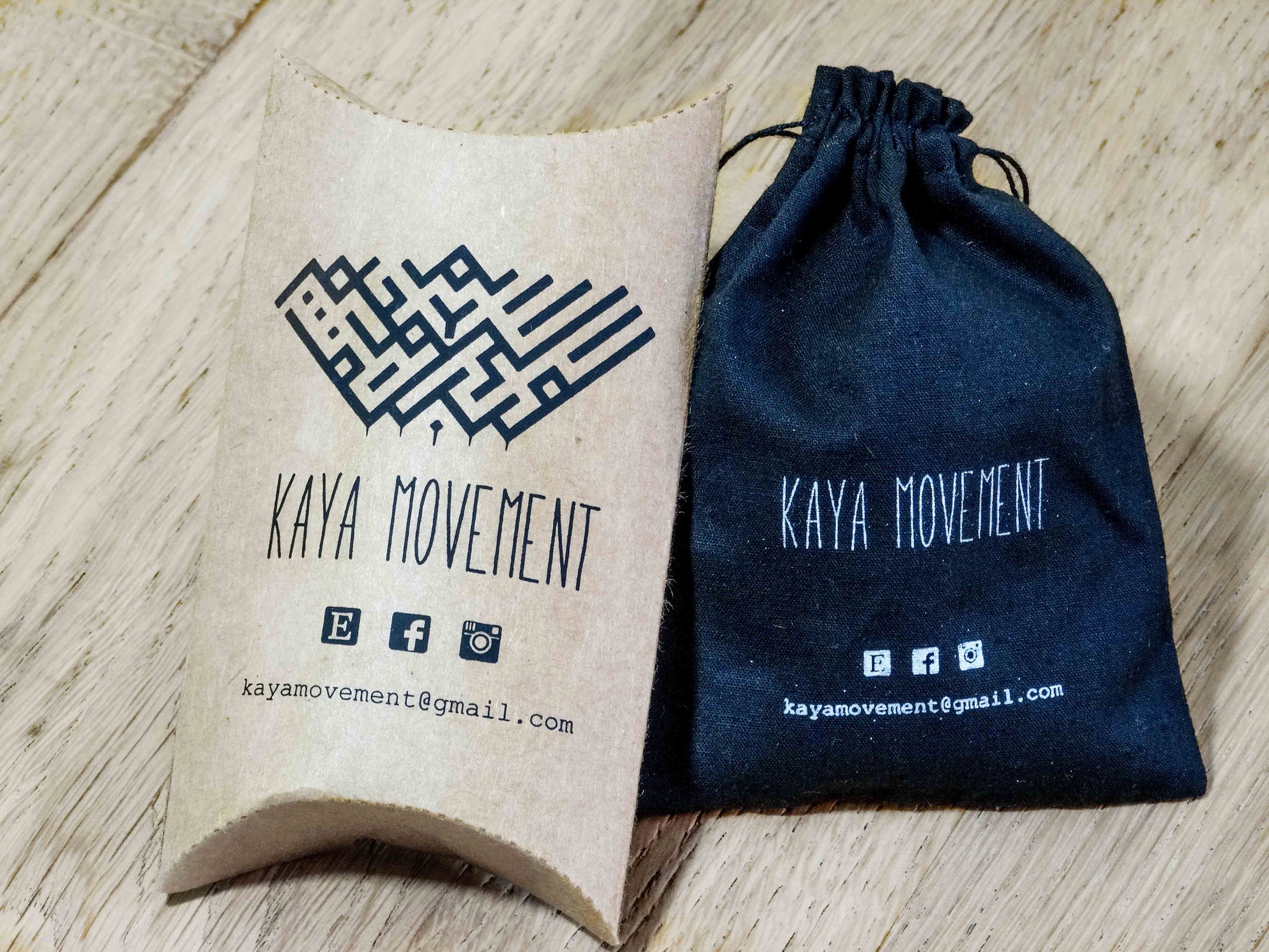 A photo showing two packaging items from Kaya Movement: a brown, pillow-shaped cardboard box and a black drawstring pouch. Both feature the Kaya Movement logo, contact email, and social media icons for Facebook, Instagram, and Twitter. These packaging options are perfect for housing your body jewelry like the SAYL Sayagata Disc Ear Weights in Gold (00 gauge).
